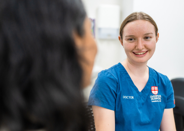 A doctor in blue scrubs smiles and listens while speaking with another person in a clinical setting, showing a calm, respectful conversation. A doctor in blue scrubs smiles and listens while speaking with another person in a clinical setting, showing a calm, respectful conversation.